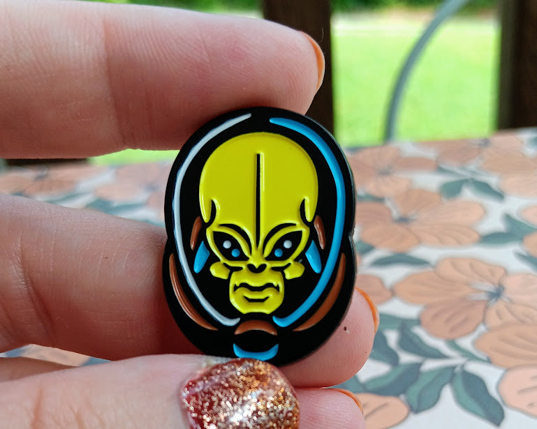Hand holding a round badge with a colorful alien design against a floral tablecloth background