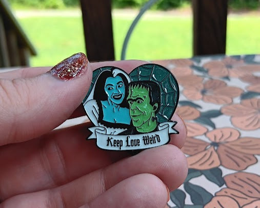 Pin with a design of a man and woman holding hands, with text 'Keep Love Weird', held by a hand against a floral-patterned surface.