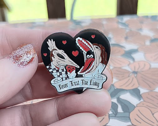 Heart-shaped pin with cartoon characters and text held by a hand on a floral surface.